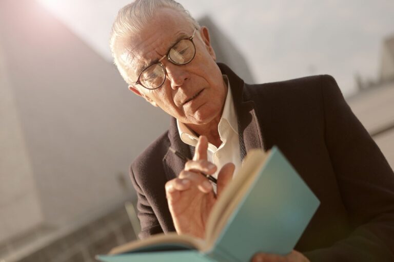Elderly man reading a book outdoors in a sunny setting, wearing glasses.